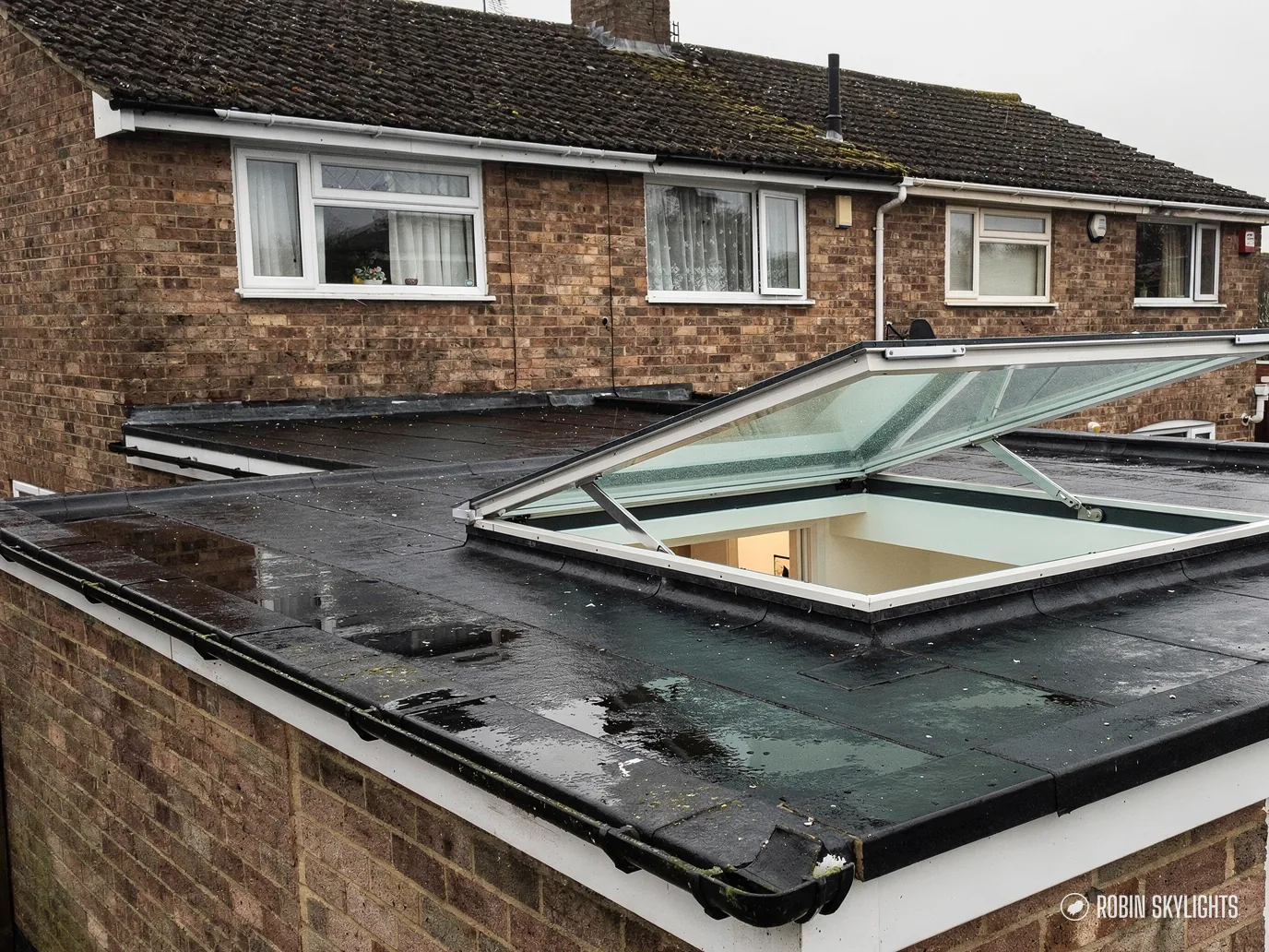 Opening flat rooflight propped open on a felted flat roof behind a brick house
