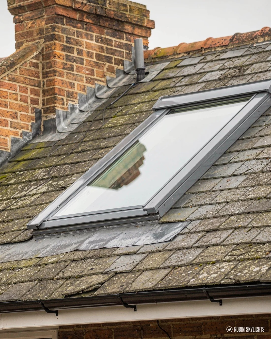 Roof window in a mossy slate roof beside a brick chimney