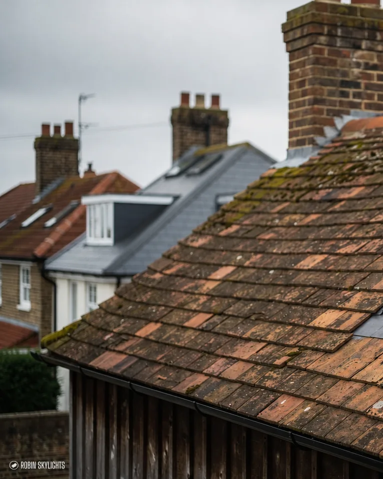 Roof window in a mossy tiled roof with chimneys behind